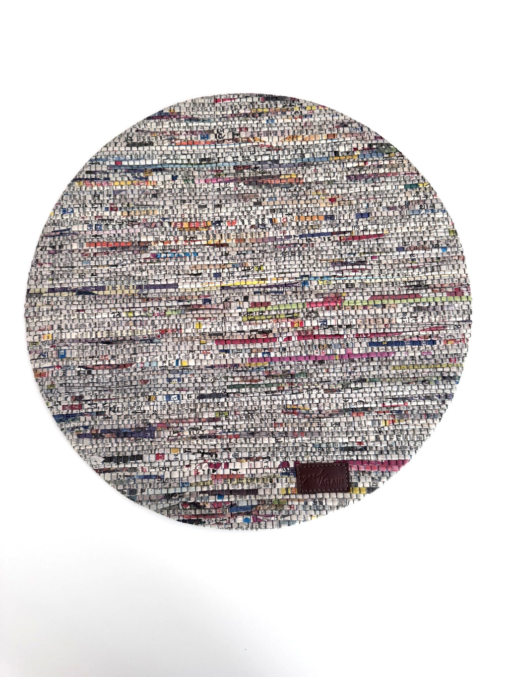 A round woven rug made from mixed-color paper strips, featuring a dense multicolor pattern with white, gray, black, red, blue, and yellow threads, finished with a small dark brown leather patch near the bottom right edge.