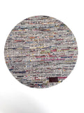 A round woven rug made from mixed-color paper strips, featuring a dense multicolor pattern with white, gray, black, red, blue, and yellow threads, finished with a small dark brown leather patch near the bottom right edge.