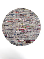 A round woven rug made from mixed-color paper strips, featuring a dense multicolor pattern with white, gray, black, red, blue, and yellow threads, finished with a small dark brown leather patch near the bottom right edge.