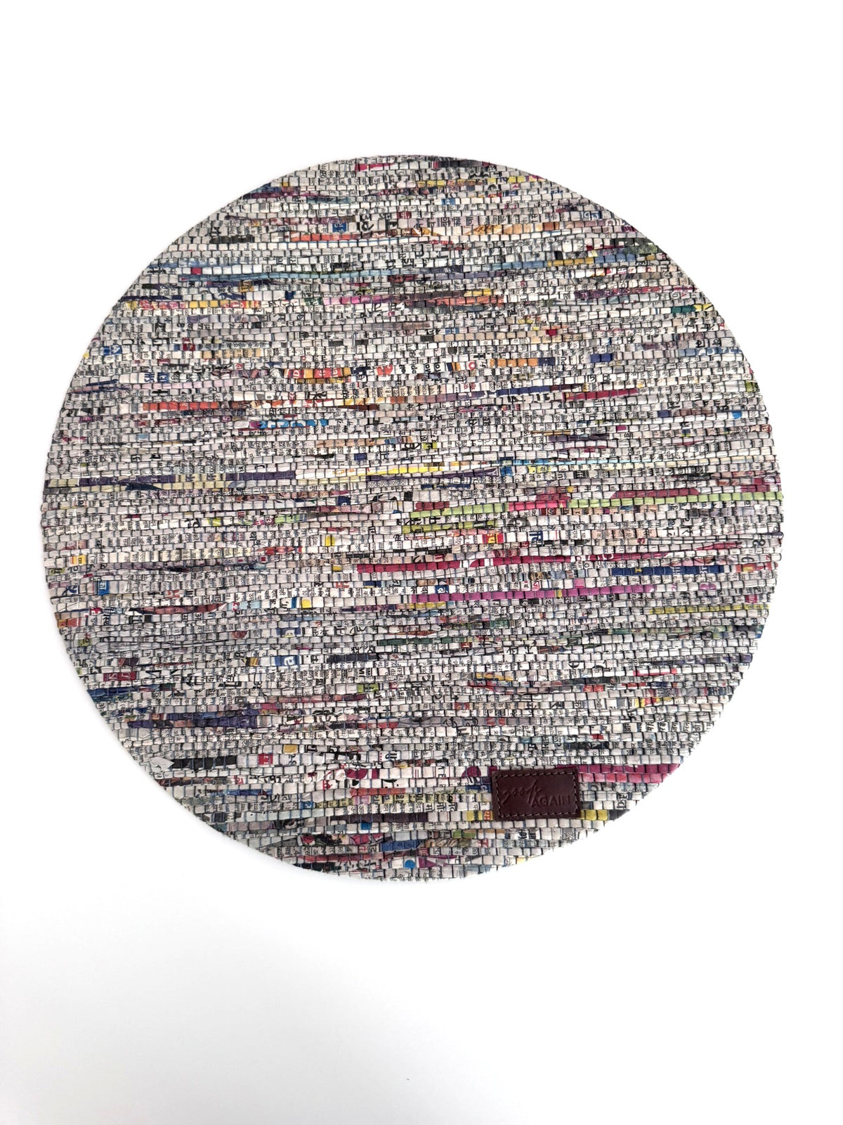 A round woven rug made from mixed-color paper strips, featuring a dense multicolor pattern with white, gray, black, red, blue, and yellow threads, finished with a small dark brown leather patch near the bottom right edge.
